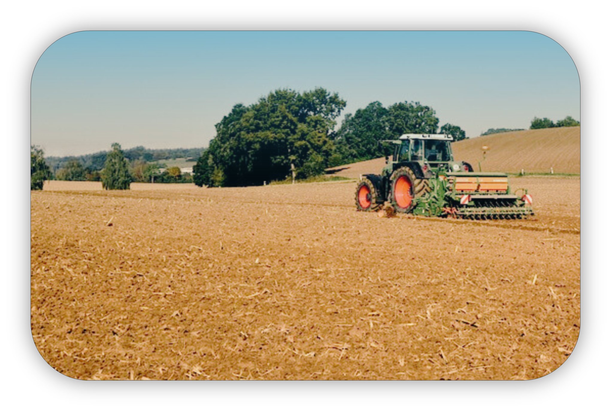 Tractor working dirt in a field with trees and a blue sky in the distance.